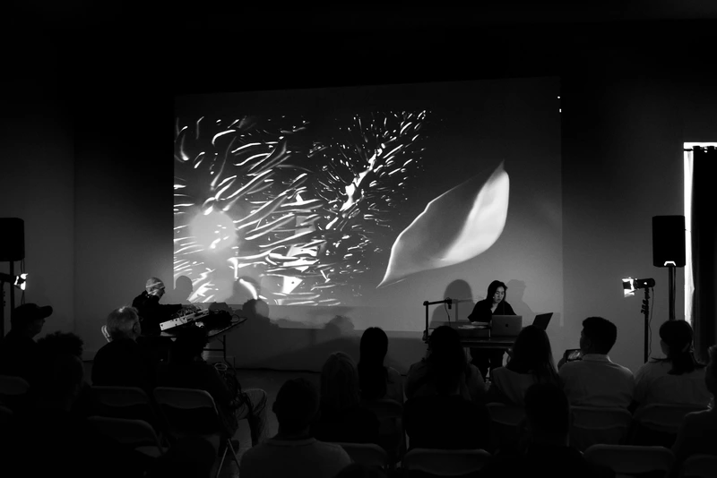 black and white image of two performers before an audience, with a projected digital image above