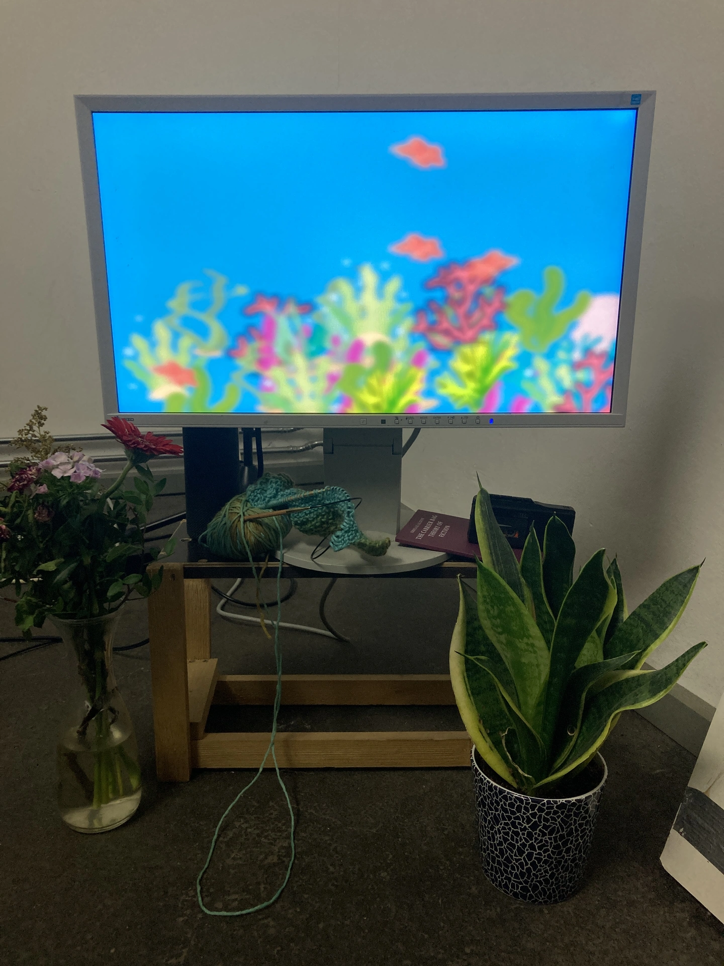 A monitor displaying a digital acquarium created in code, framed by a plant and flower bouquet, yarn, microcassette and book A monitor displaying a digital acquarium framed by a plant and flower bouqet, along with knitting needles and yarn, microcassette recorder and a book