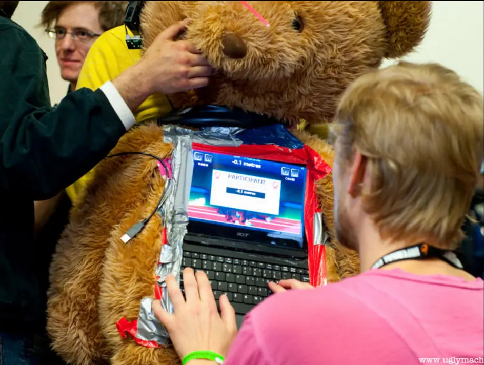 A giant stuffed animal with a computer duct taped to its stomach is the host of a video game, played by someone whose back is turned to the camera