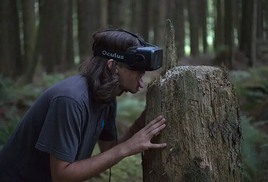 A person wearing a VR headset, tongue extended, about to lick a stump in the forest.