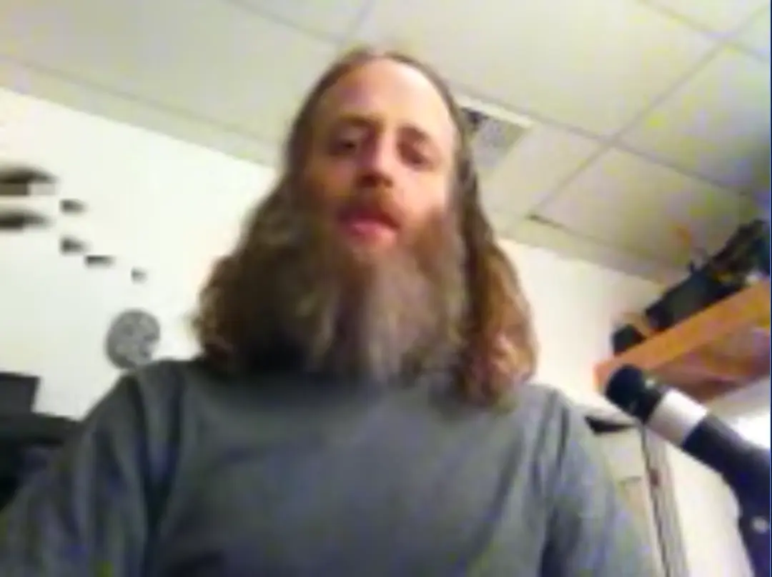 Guru Rugu with beautiful bushy beard looks into the camera, a mic off center, about to lead a meditation over the radio.