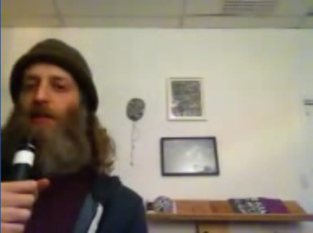Guru Rugu with bushy beard sits on the left side of the frame, holding a microphone and speaking to his radio audience.