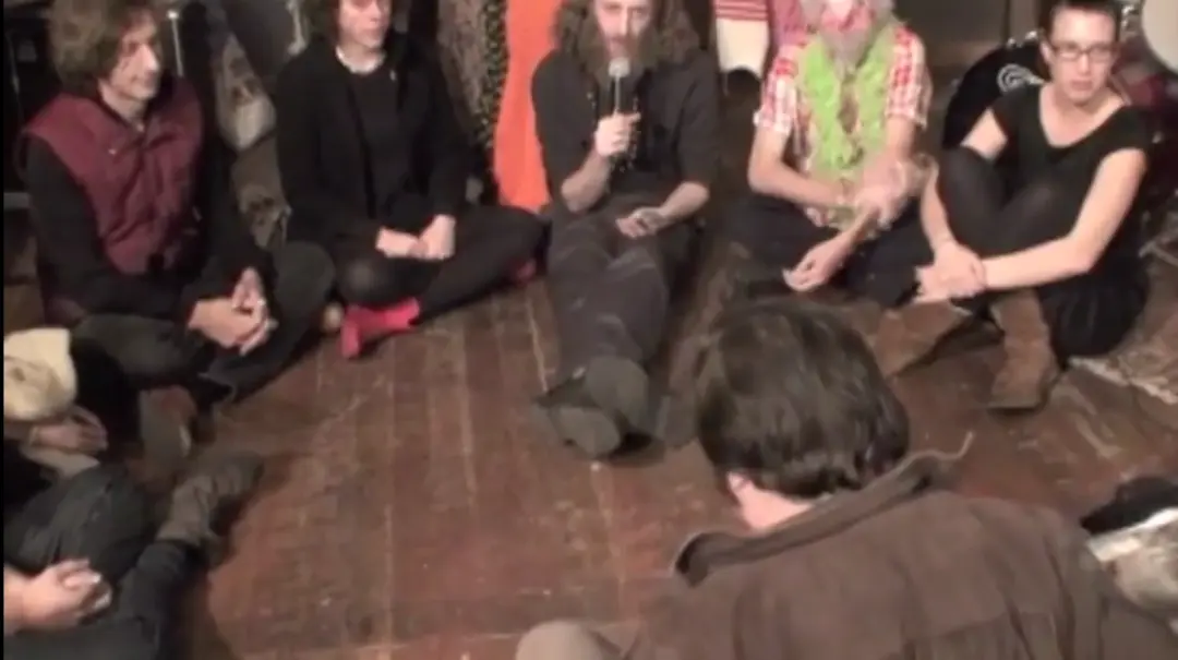 A group of people sit on the floor in a magic circle around Guru Rugu, who holds a microphone.