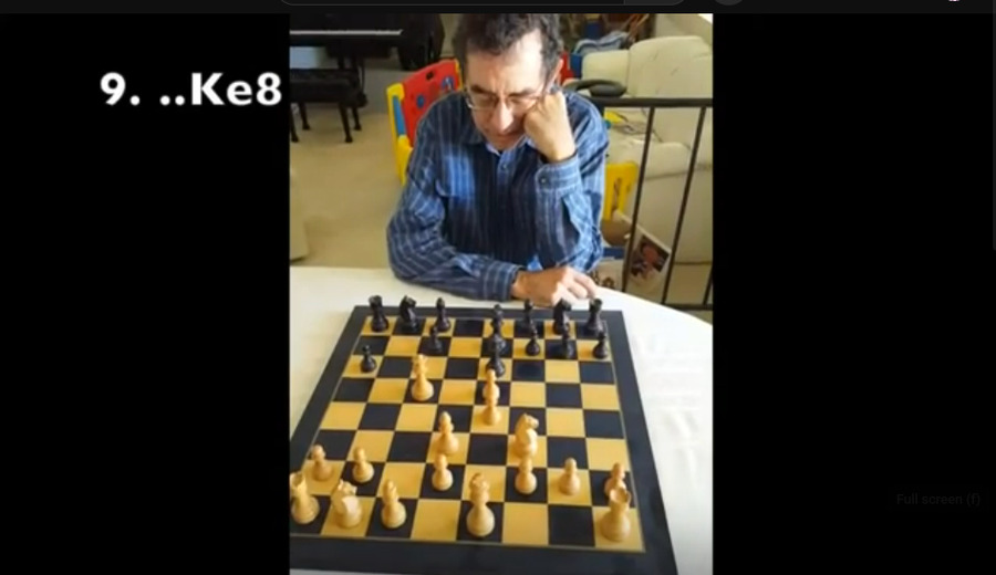 An older man sits in a contemplative mood looking at a chessboard. Behind him is a living room with piano bench, kids' playpen, baby gate and couch. The chess move 9. ..Ke8 in white text on top.