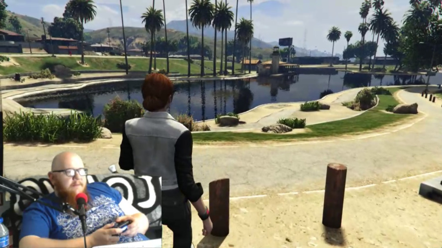 A screenshot of a balding man reclining, speaking into a microphone with orange cover, embedded on top of a live screenshot of a videogame (Grand Theft Auto V) with their character's back to us, looking at a virtual pond with palm trees surrounding.