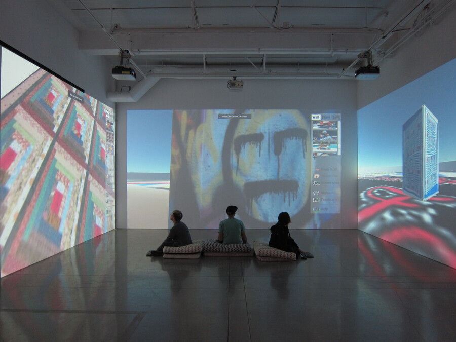 Installation view of Messlife. Three people sit on cushions in the center of the gallery looking at three screens. On the left projection is a floor quilt. In the center is a wall with a graffitti face. On the rice is a block with a quilt on an abstract ground.