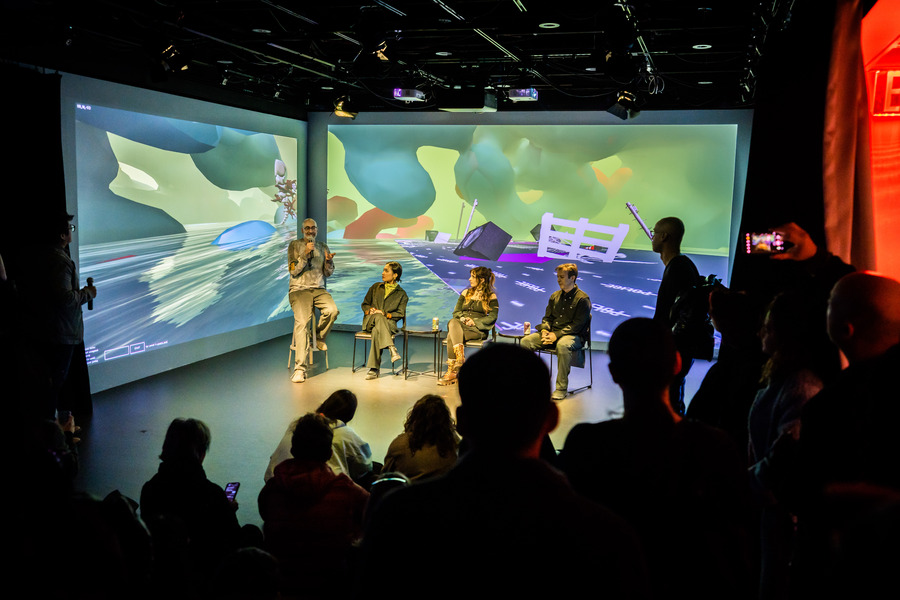 A group of 4 people sit in front of an audience in a darkened room in front of a dual screen projection, showing a green atmosphere and the floor from Messlife