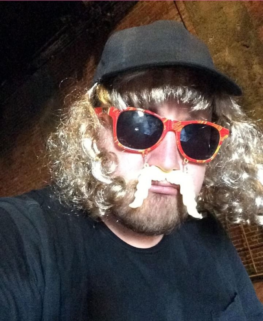 DJ Bearings wears a disguise with blond curly wig, black hat, red sunglasses and plastic mustache.