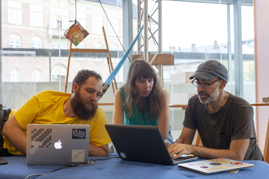 Three people sitting behind two laptops. Lee in a blue patterned hat with glasses and white beard is typing while two other people look on.
