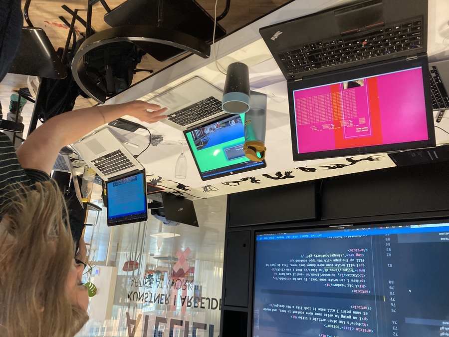 A woman looks at a table of laptops showing creatively designed websites, in front of a larger monitor showing HTML code. The laptop screen on the left shows pink background with ASCII art. In the middle a green background with large photos.