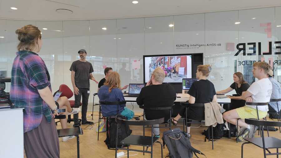 A group of people in a workshop sitting around a larger monitor. Lee stands in the front left of the room, speaking.